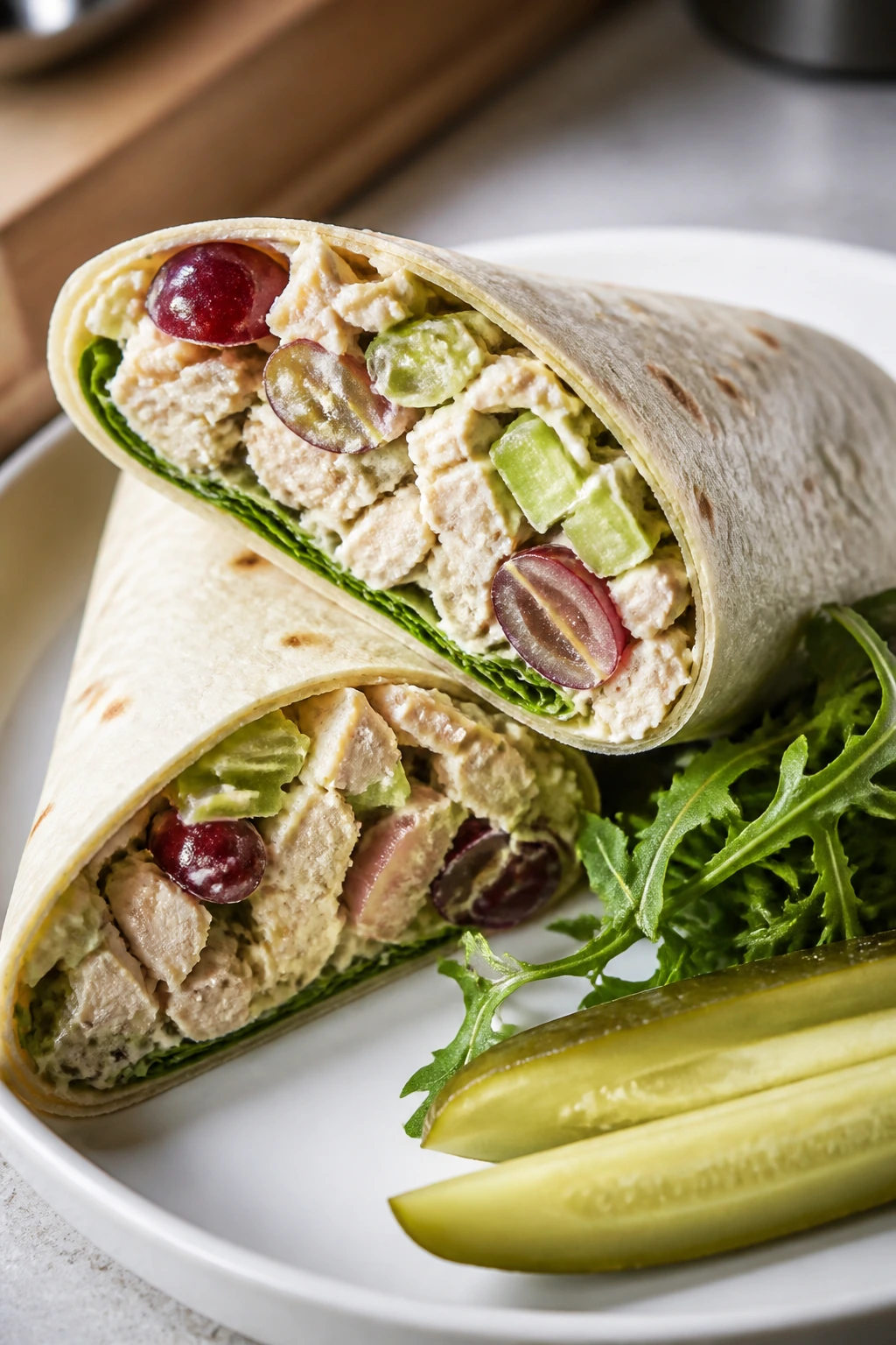 Close-up of diagonally sliced chicken salad wrap showing crisp celery and grapes on a plate with greens and pickles