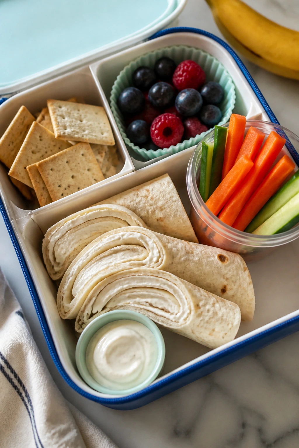 Lunchbox showing tortilla with cream cheese swipe and carrot sticks with dip