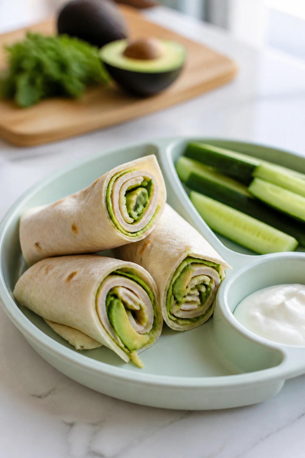 Lunch plate featuring turkey avocado roll-ups and cucumber spears on a sunny counter.