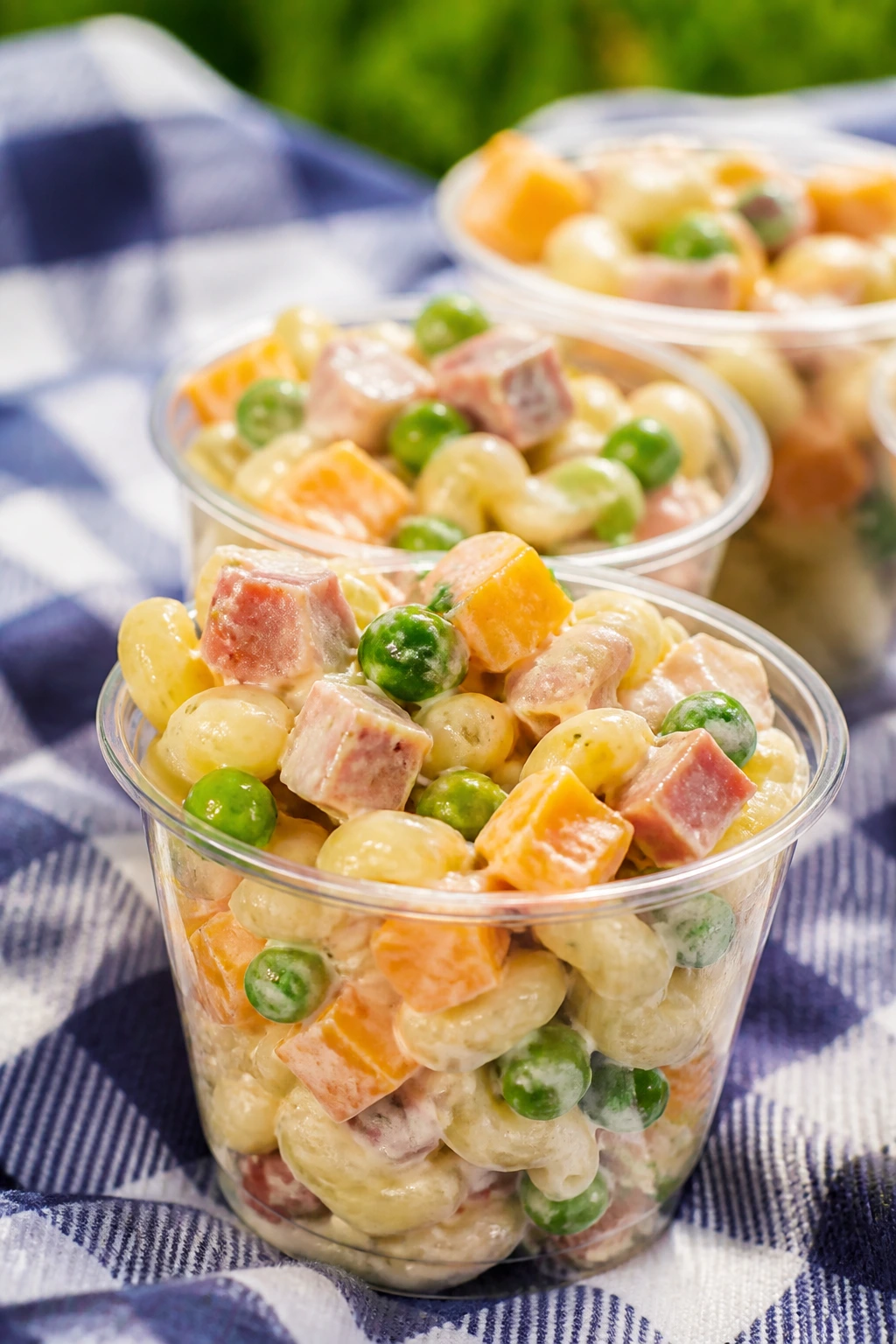 Small cups of pasta salad with peas and ham on a picnic blanket.