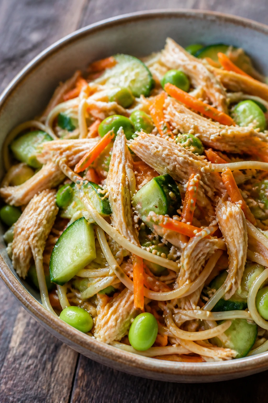 Close-up of sesame chicken noodle salad with noodles, chicken, carrots, cucumber, and edamame in a light dressing