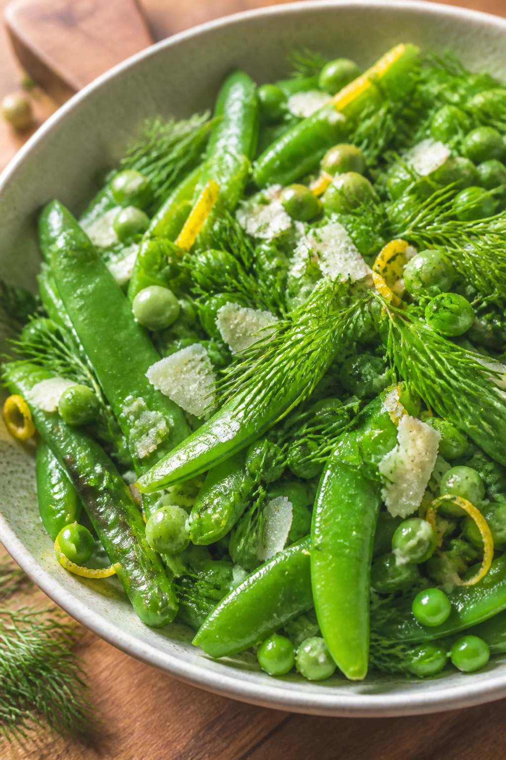 Close-up of lemon-dill snap pea salad in a bowl with bright green peas and Parmesan on a wooden counter