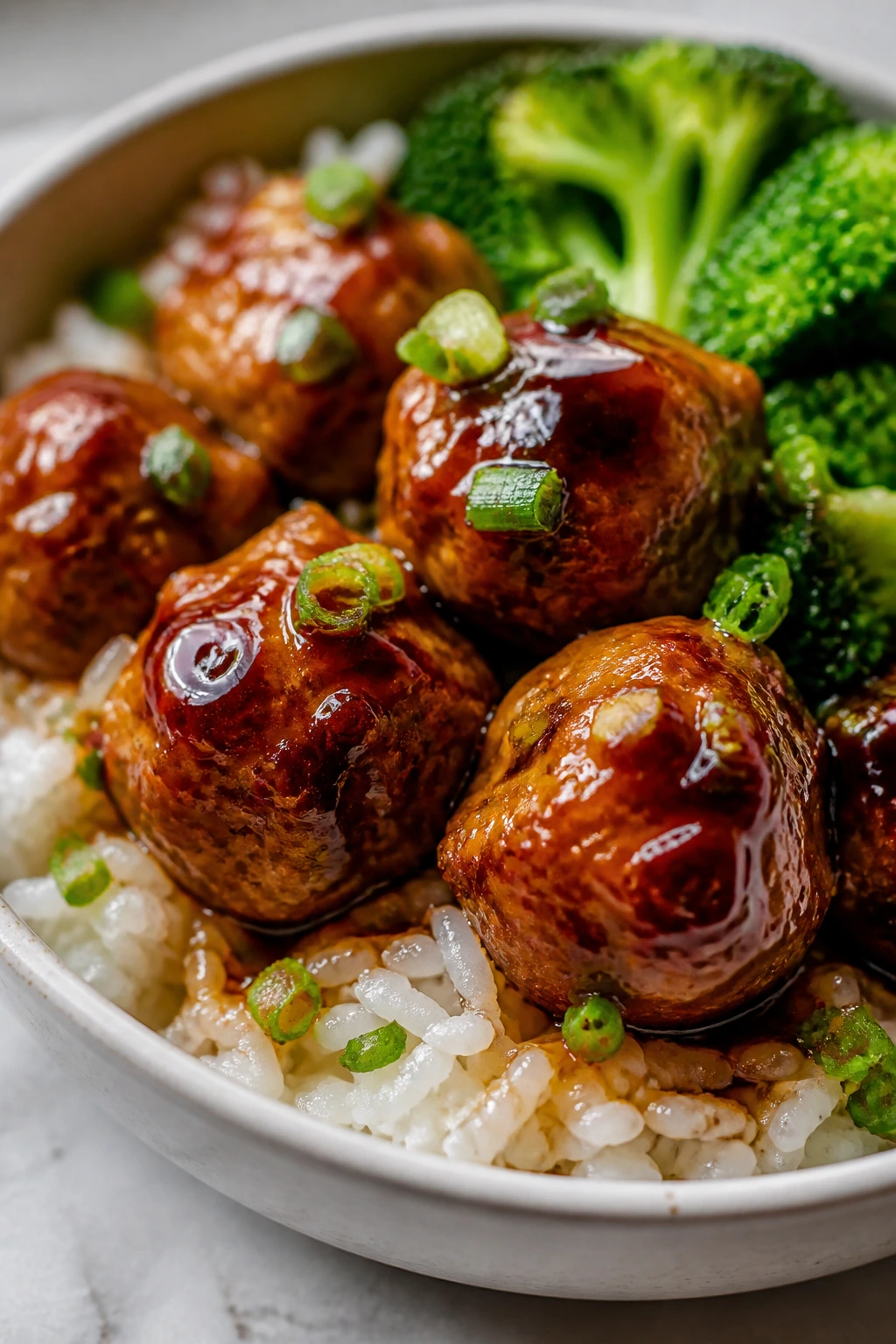 Glossy teriyaki meatball on rice with broccoli in a bowl, kitchen setting.