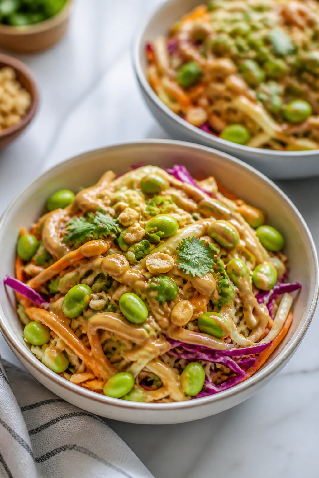 Two bowls of Thai peanut slaw with noodles and edamame in a peanut sauce on a bright kitchen counter.