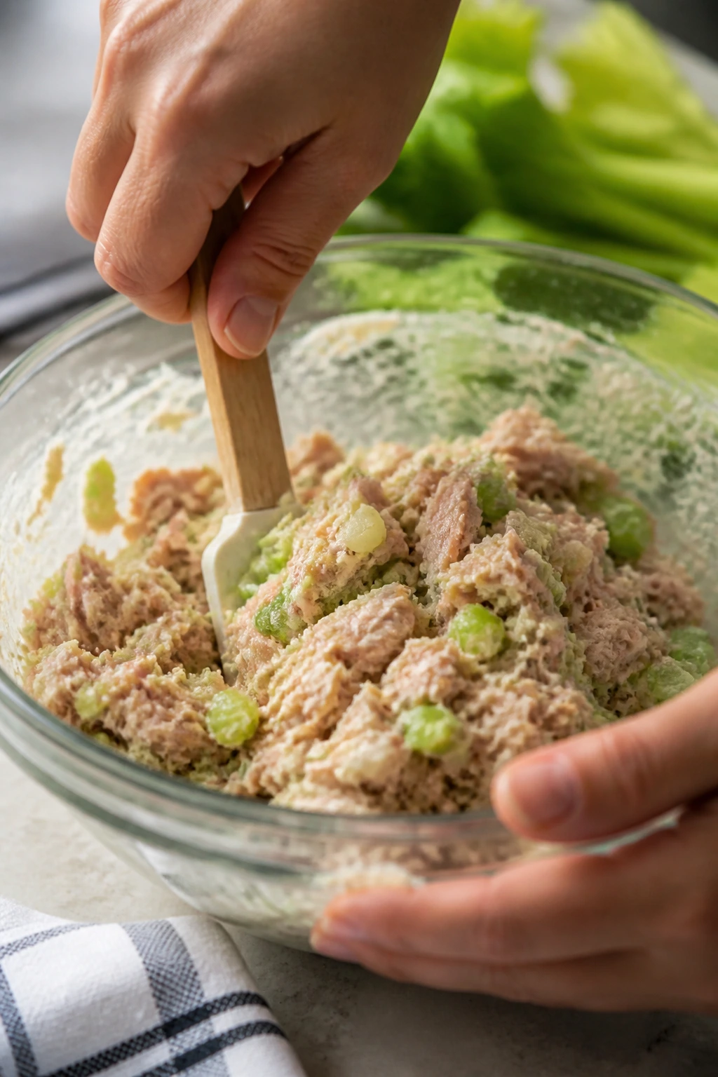 Hands folding tuna salad with celery and onion in a glass bowl in a kitchen